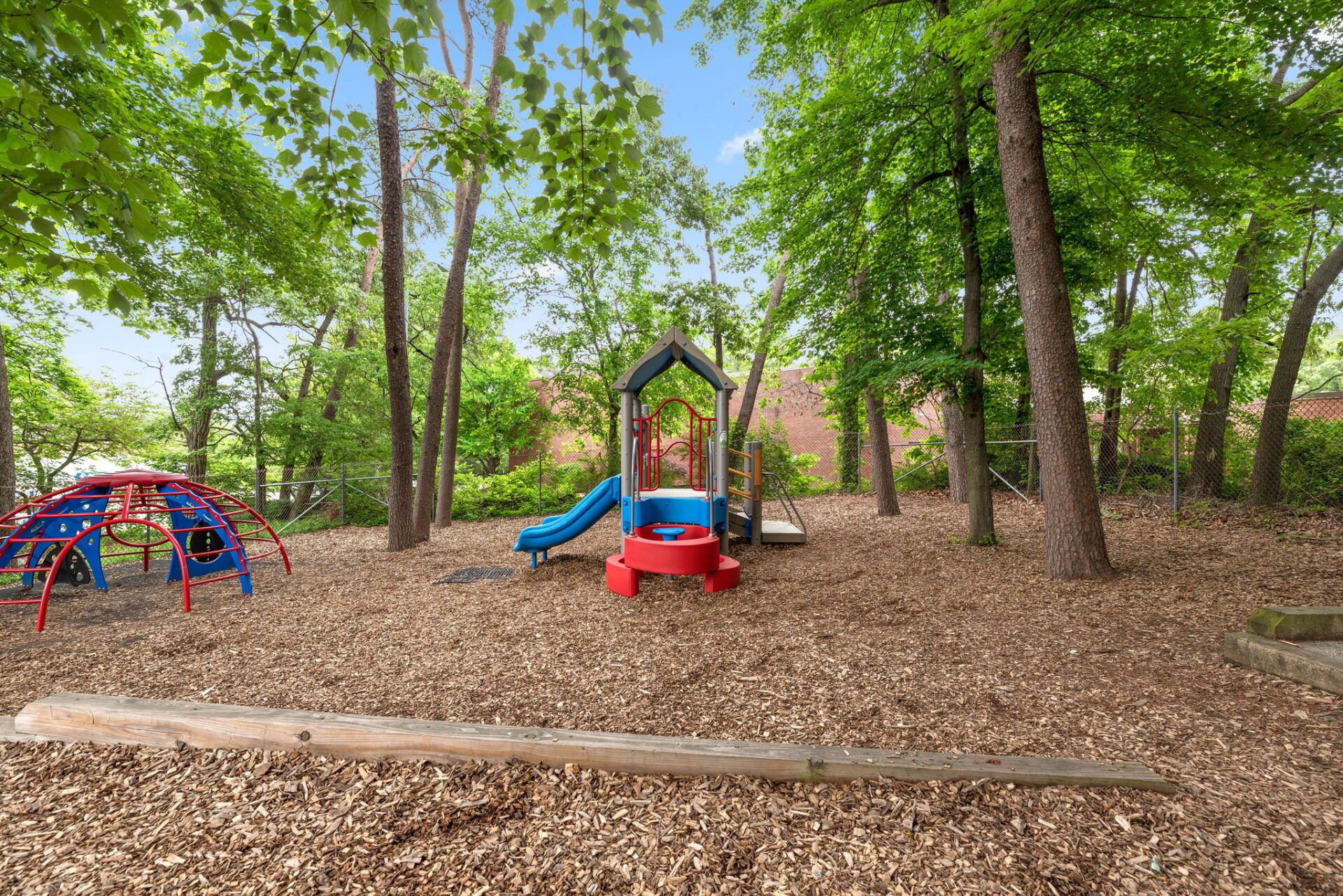 Playground in a wooded area with a slide, climbing structure, and woodchip ground cover. Trees surround the play area.