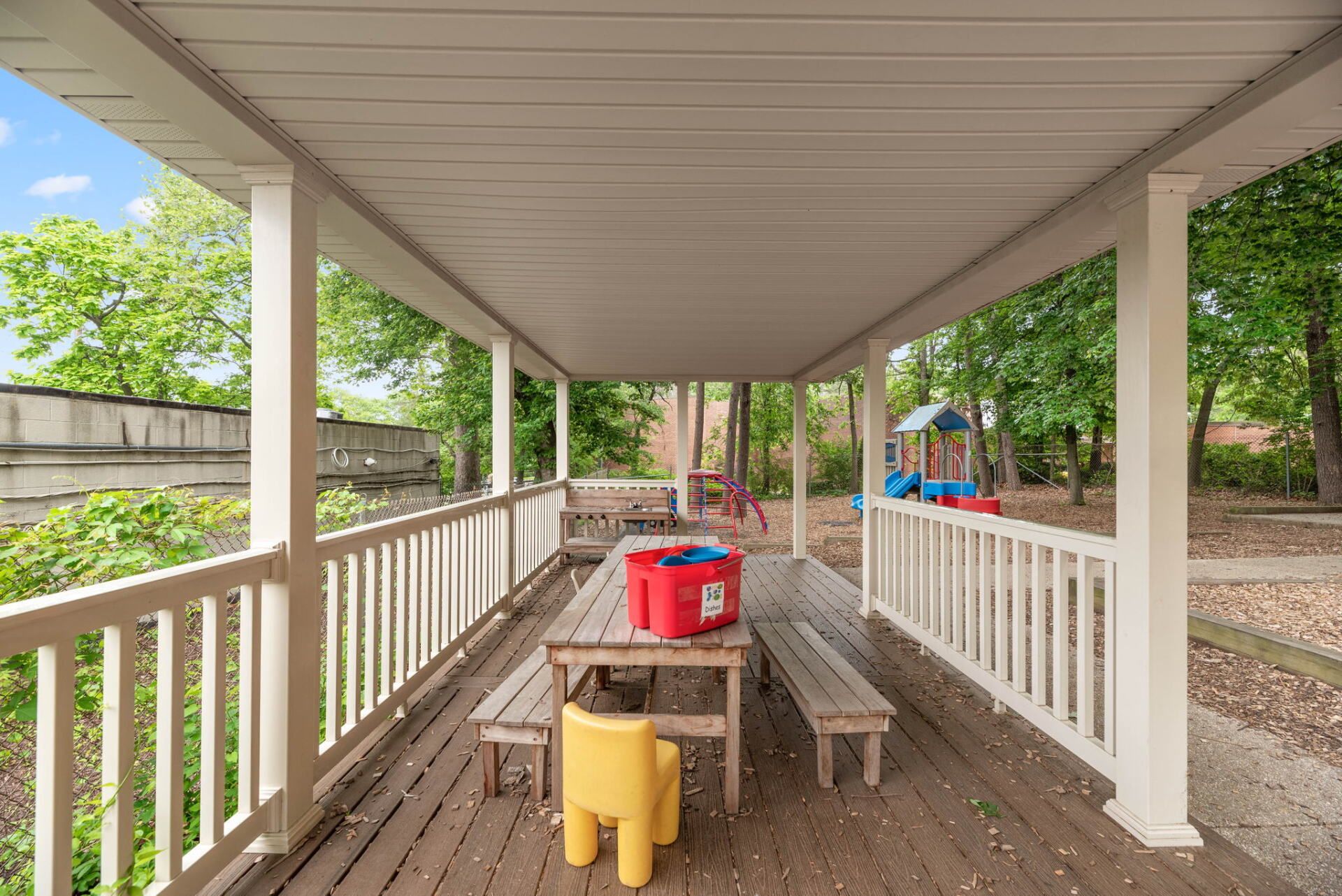 Covered outdoor deck with a table, benches, and a yellow chair; a playground and trees are visible in the background.