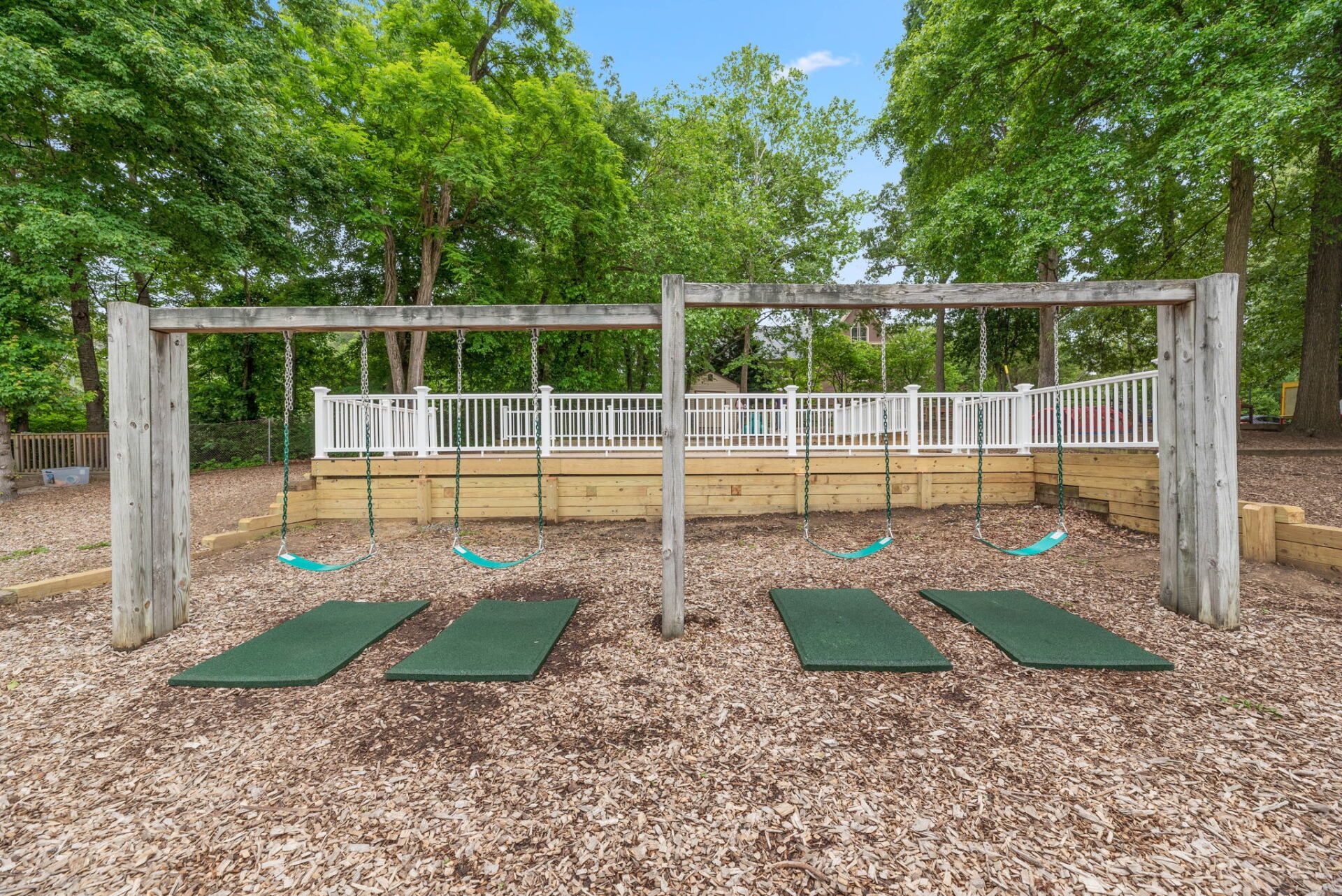 A children's playground with two swing sets and green safety mats on wood chip ground.