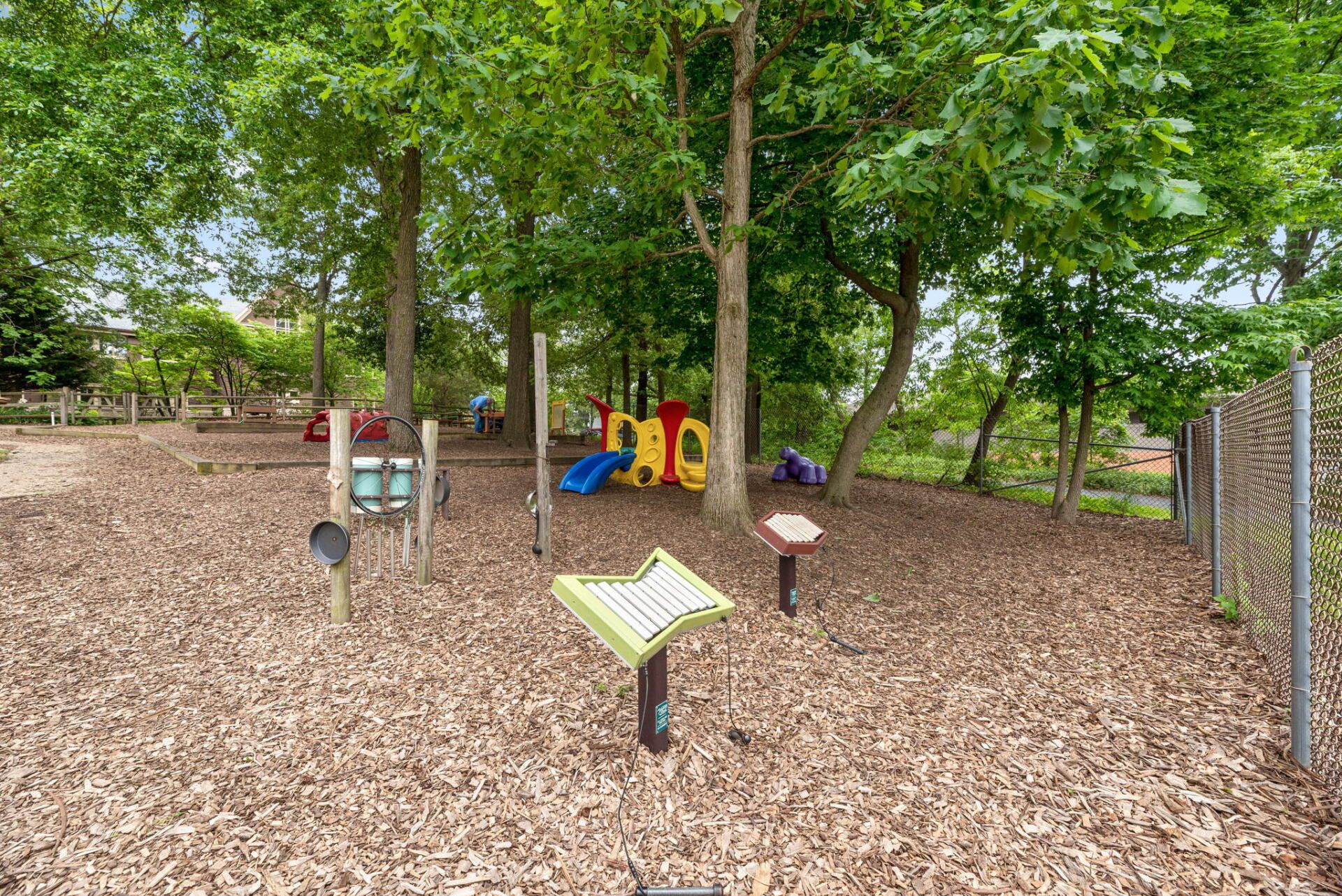 Playground with musical instruments and a small slide set under trees. Ground covered in wood chips.