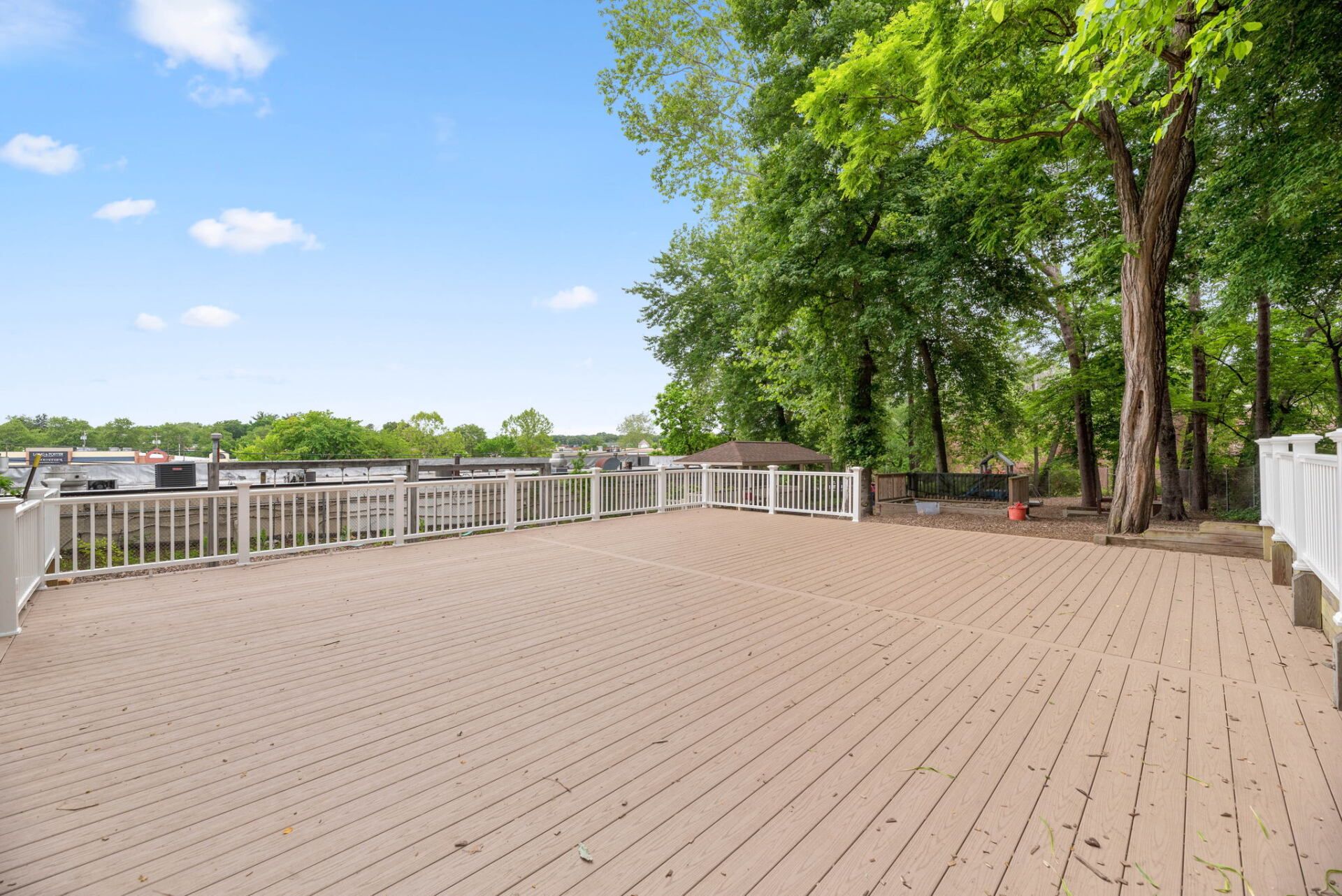 Large, empty wooden deck surrounded by a white railing and lush green trees under a blue sky.