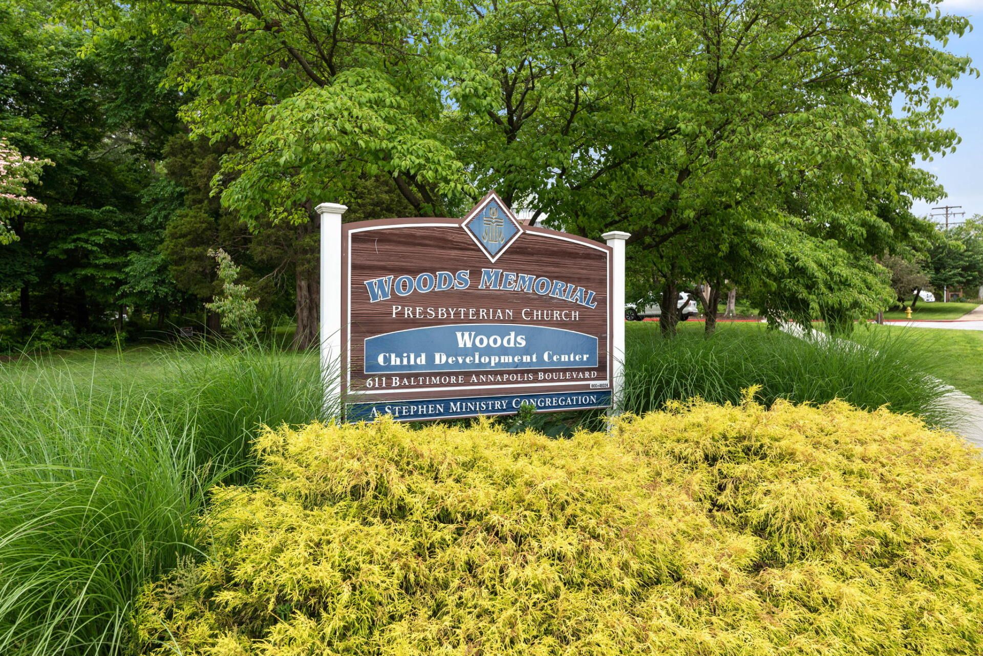 Sign for Woods subdivision, with brown and white trim, surrounded by green shrubbery and trees.