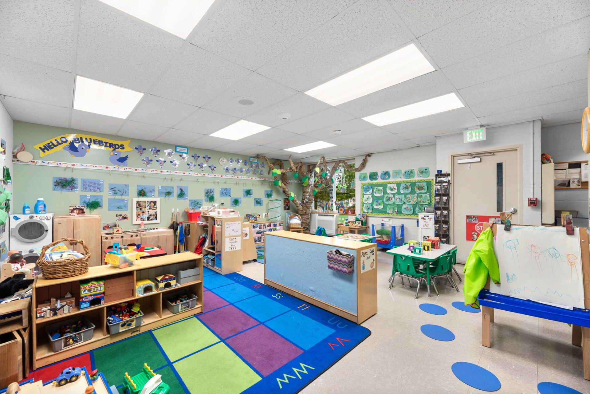 A bright, well-organized preschool classroom with colorful toys, art, and various activity stations.  Soft blue and green tones create a welcoming environment.