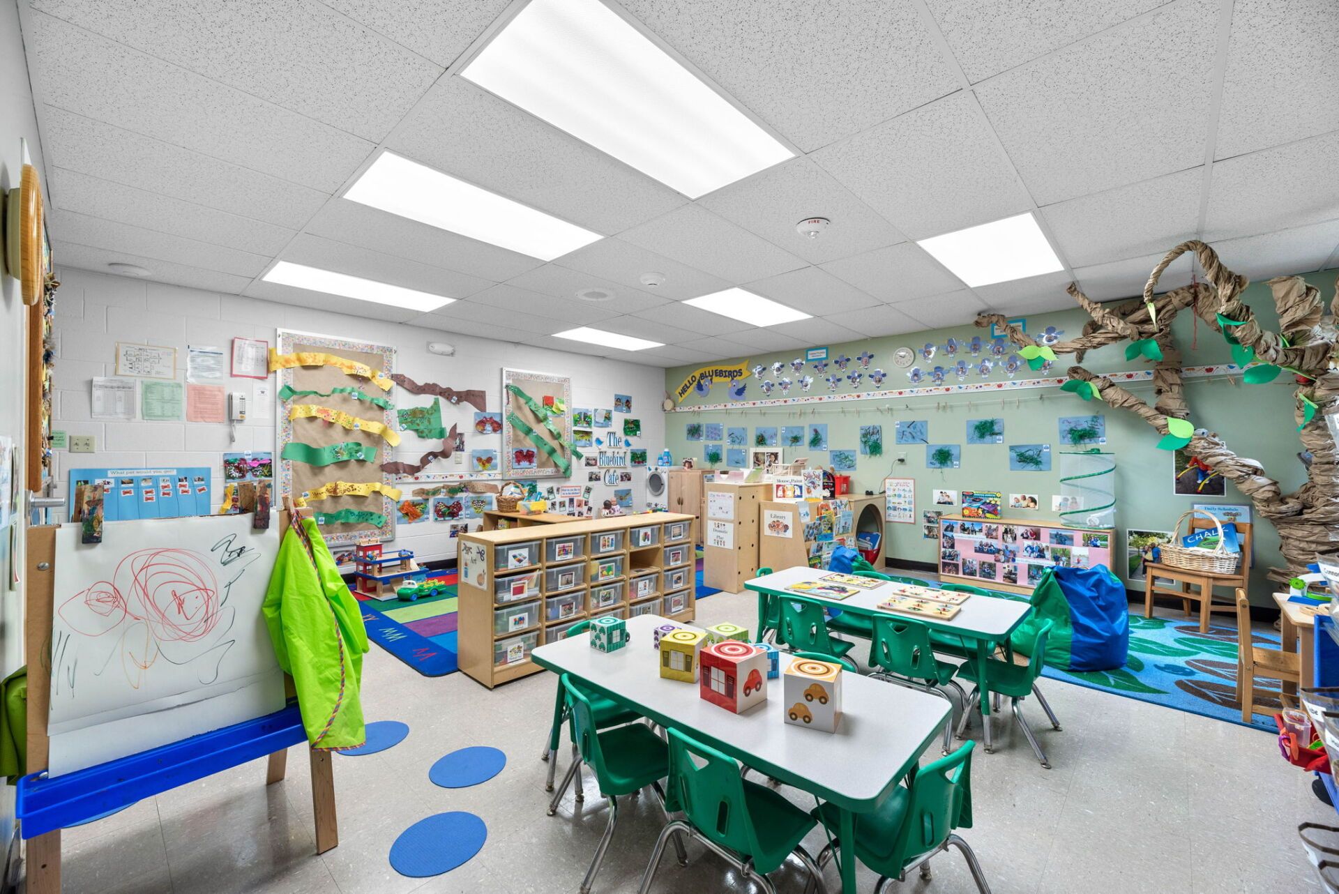 A bright preschool classroom. Tables and chairs surround a central area with art projects and learning materials.
