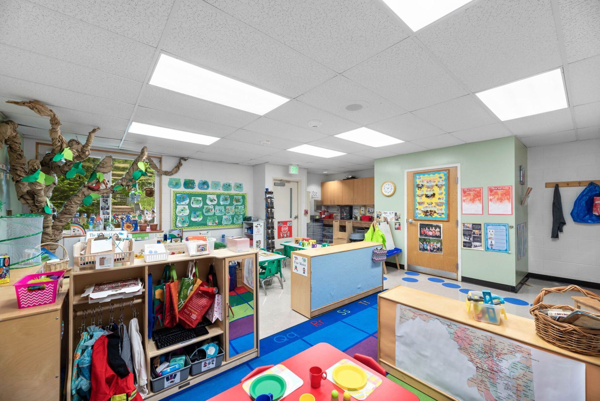 A brightly lit preschool classroom with colorful decorations, a tree-like structure, and children's materials on display.