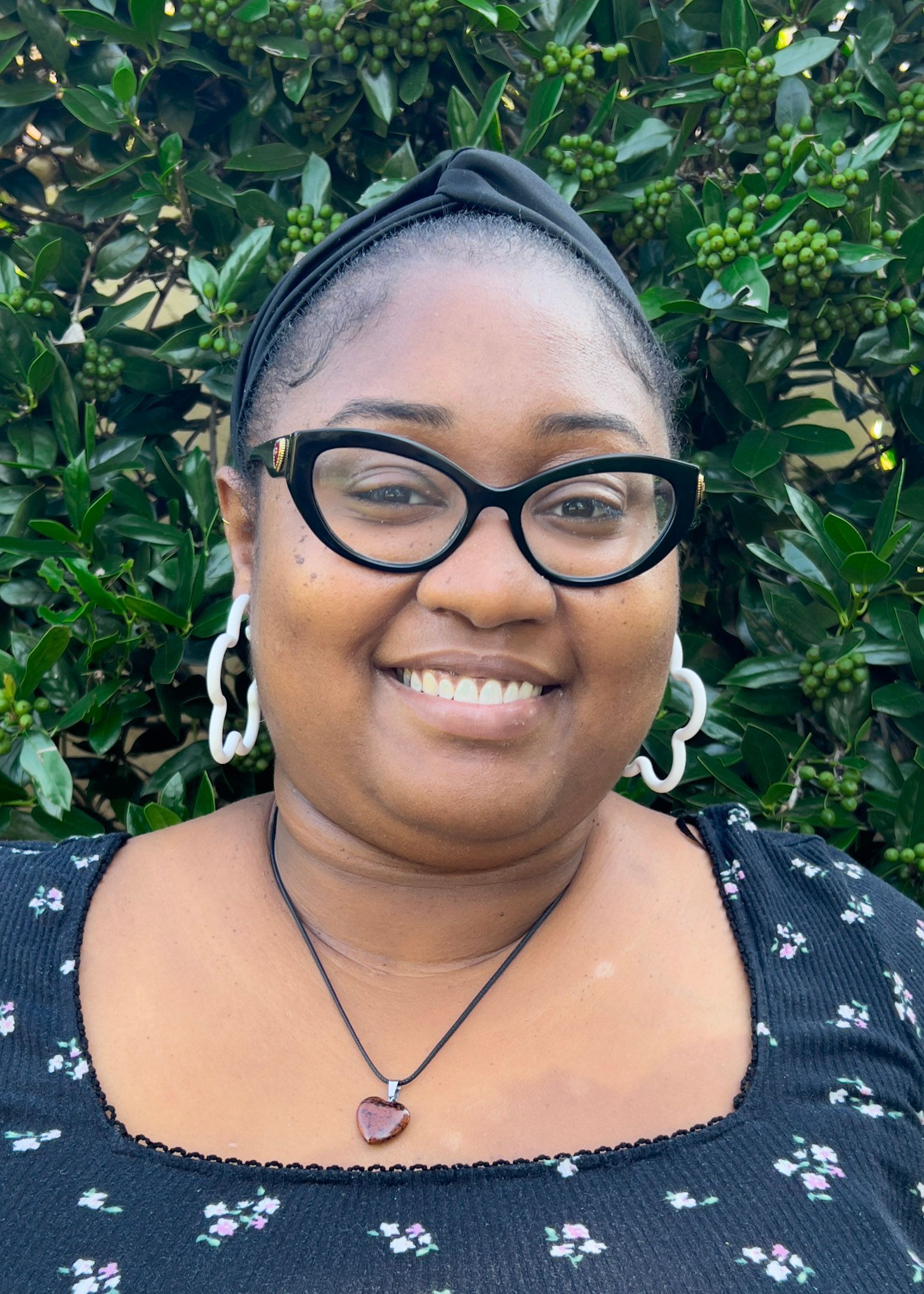 Woman with dark skin wearing glasses, smiling, outdoors, with long, dark, and auburn-streaked locs.