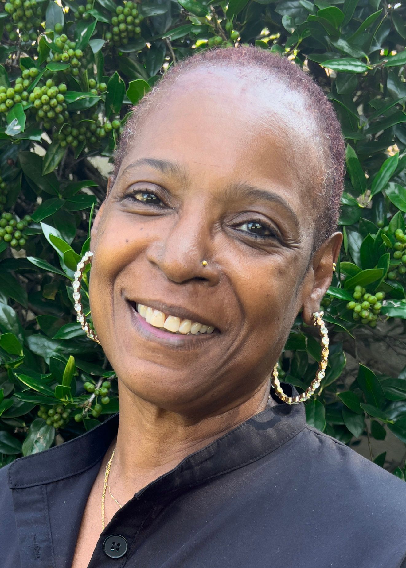 Smiling Black woman with short hair, wearing large hoop earrings and a black zip-up jacket against a blue background.