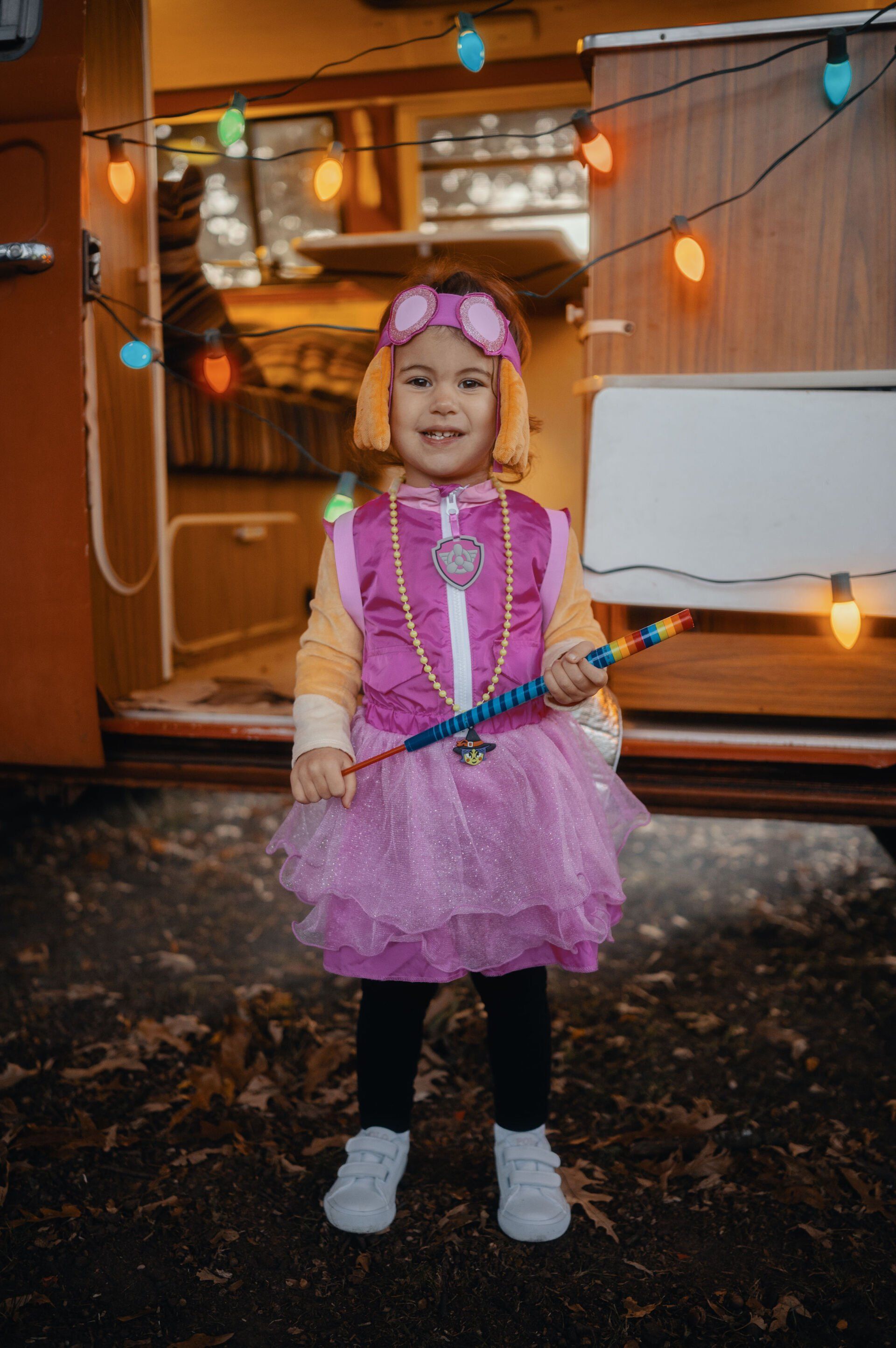 A young child dressed as Skye from Paw Patrol, in a pink and orange outfit, smiling and holding a wand, with a trailer in the background.