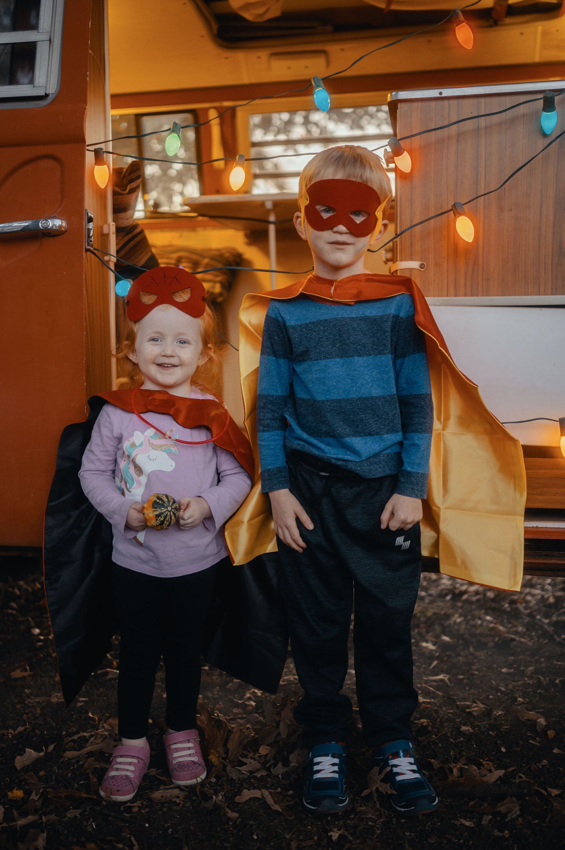 Two children dressed as superheroes, smiling, standing in front of a camper decorated with lights.