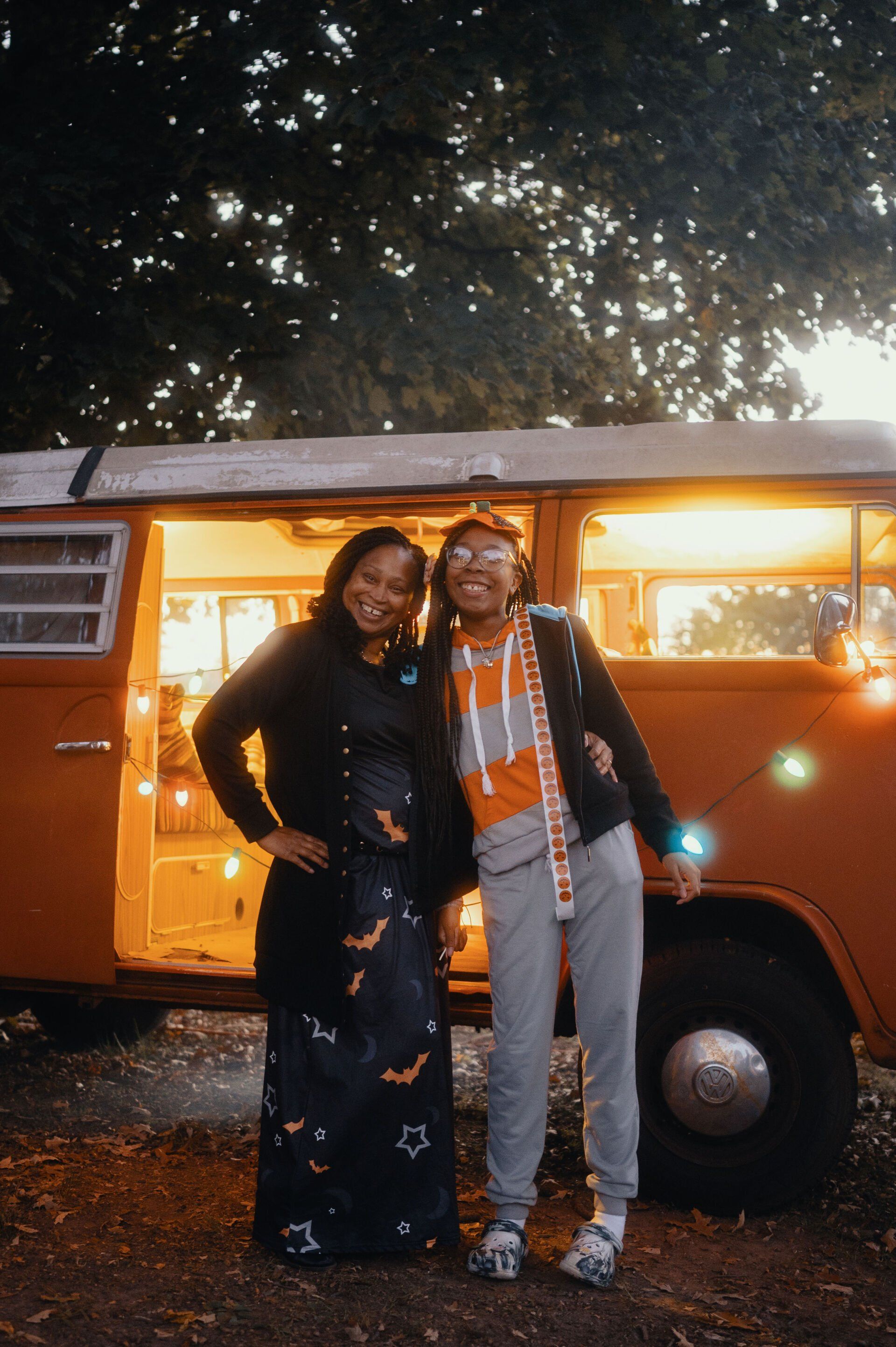 Two smiling women pose by an orange van with lights on. One wears a black dress with bats, the other, a gray sweatsuit and orange accents.