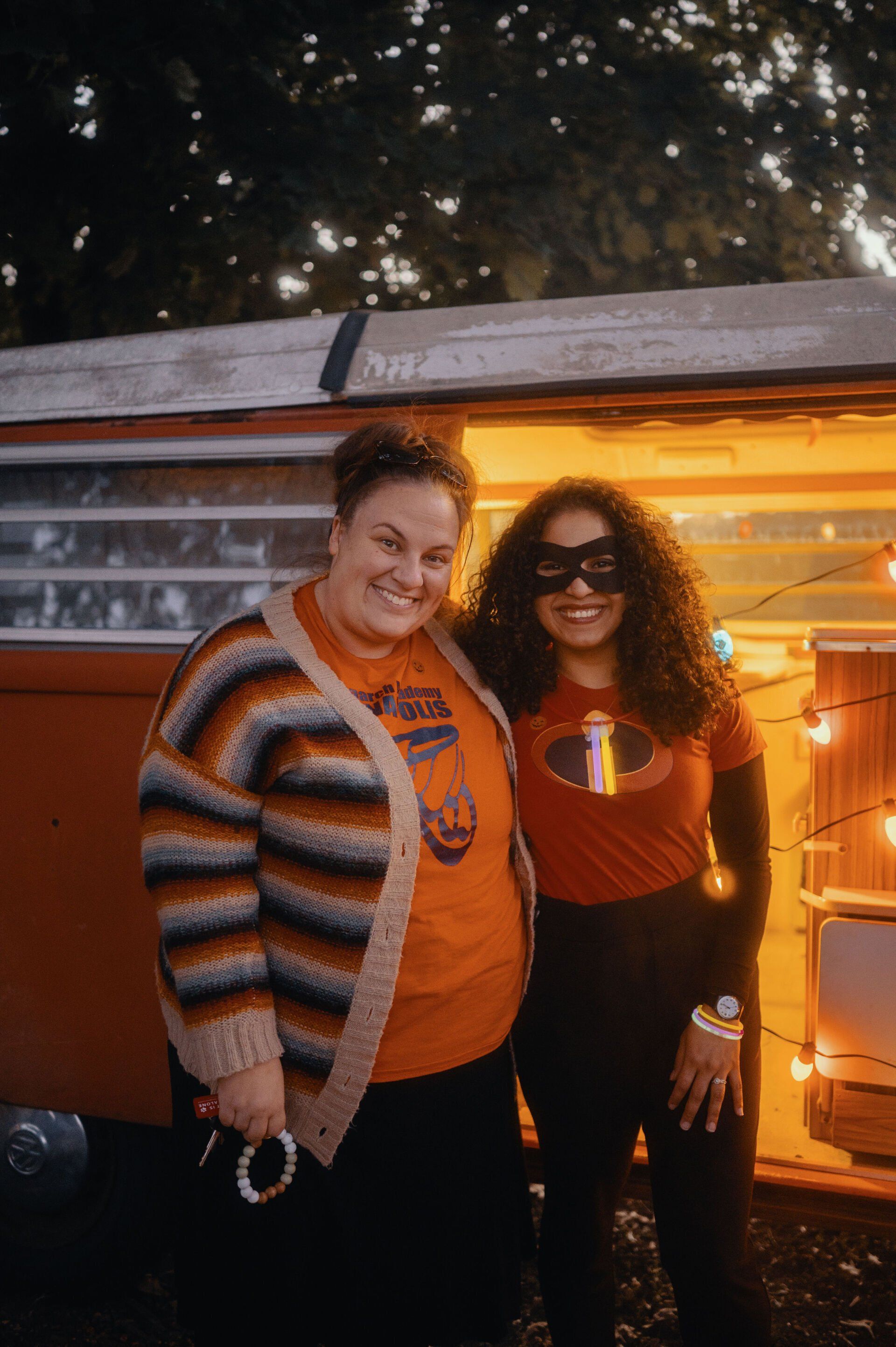 Two women in front of a rustic trailer. One wears a cardigan and a fist-themed shirt, the other is in an Incredibles costume.