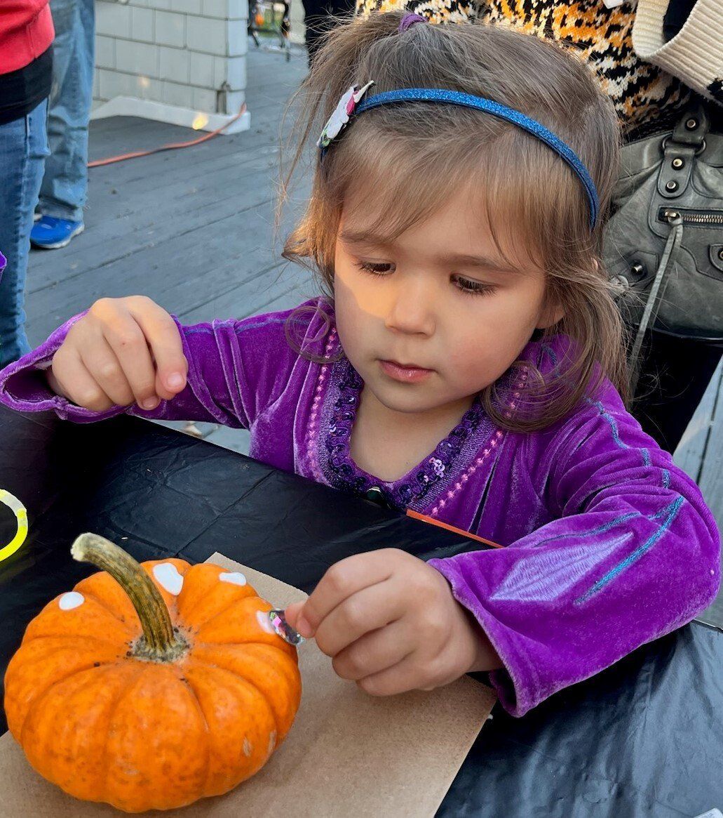 A young girl in a purple jacket decorates a small pumpkin with white dots at an outdoor table.