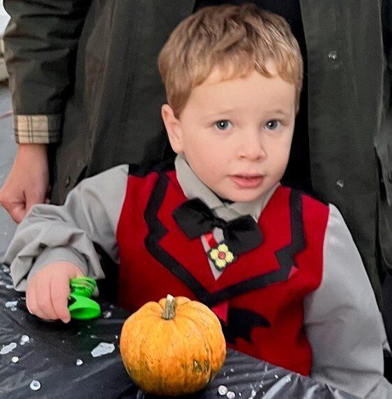 A young boy in a vampire costume poses with a small pumpkin. He's wearing a red vest and gray shirt, looking directly at the camera.