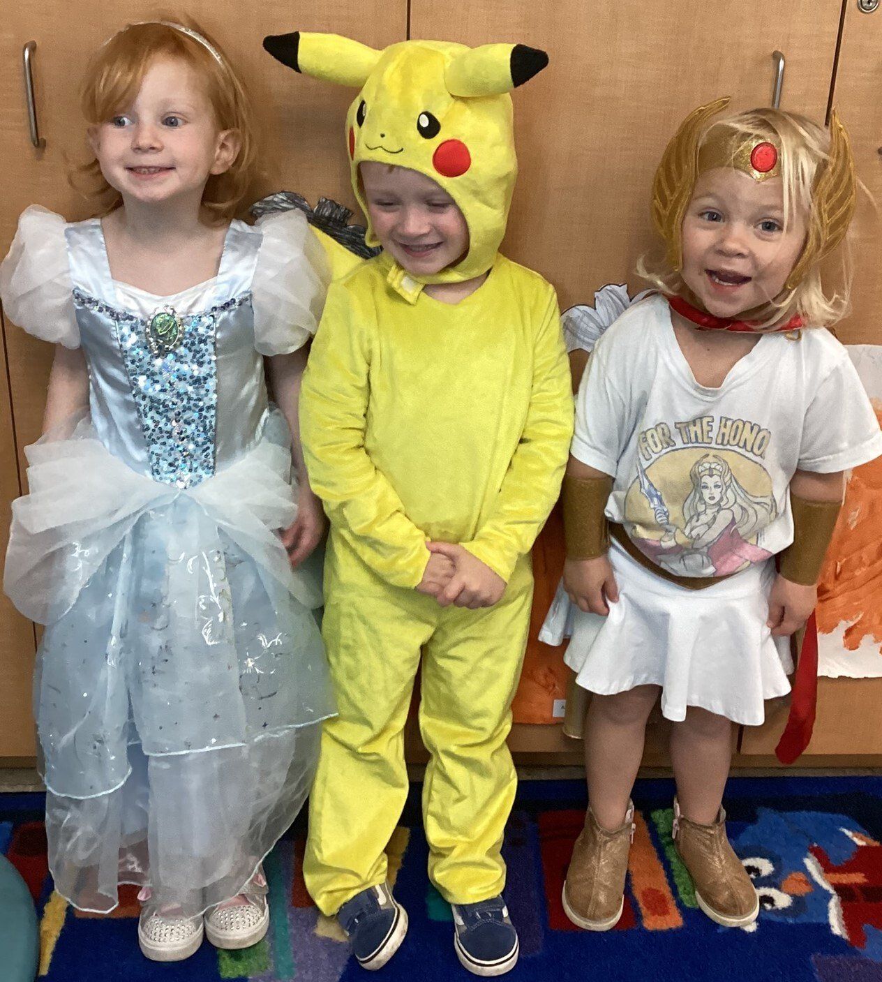 Three young children in costumes: a princess, Pikachu, and a Greek goddess, smiling in a classroom setting.
