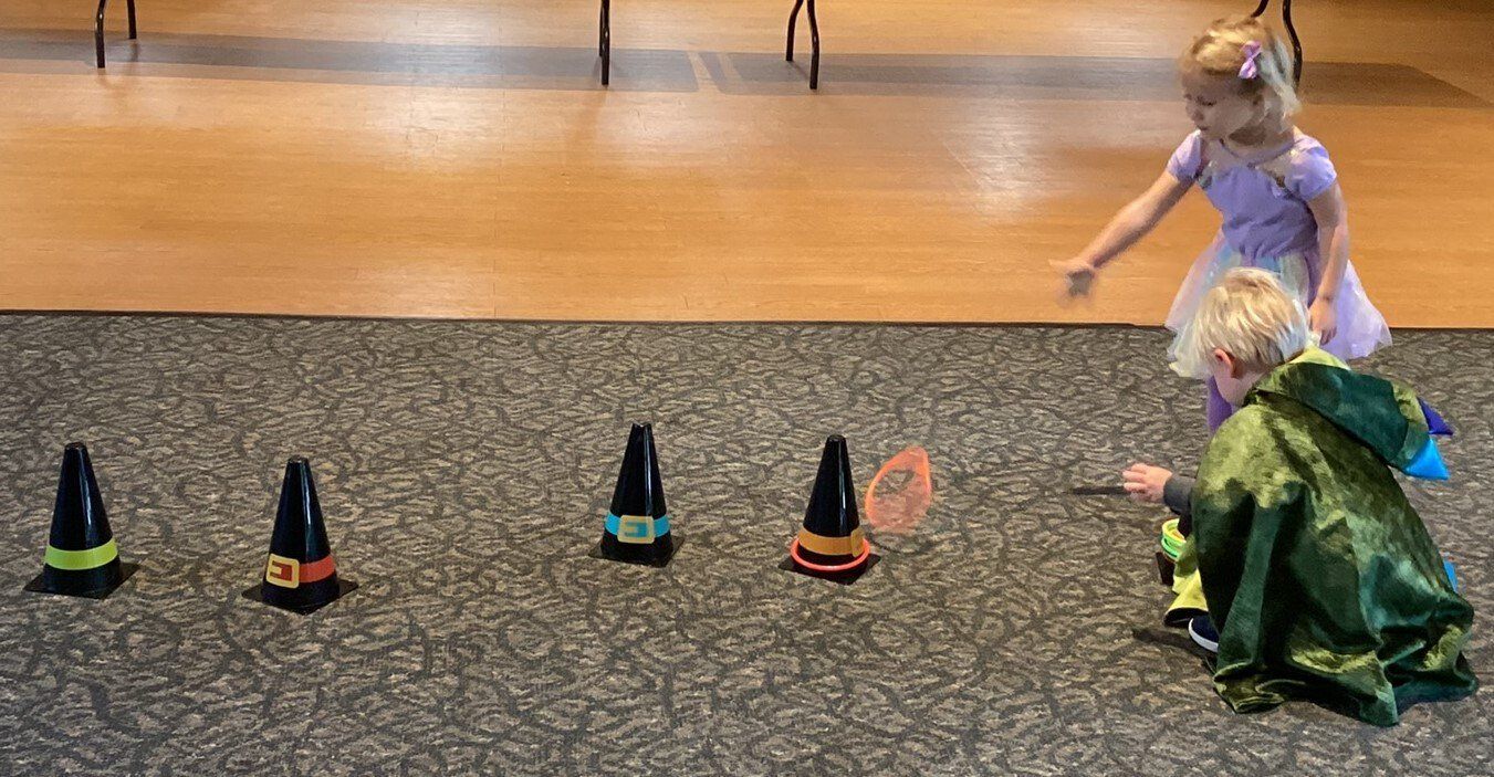 Two children, one dressed as a fairy, the other as a dragon, play a game with orange cones and a pink butterfly.