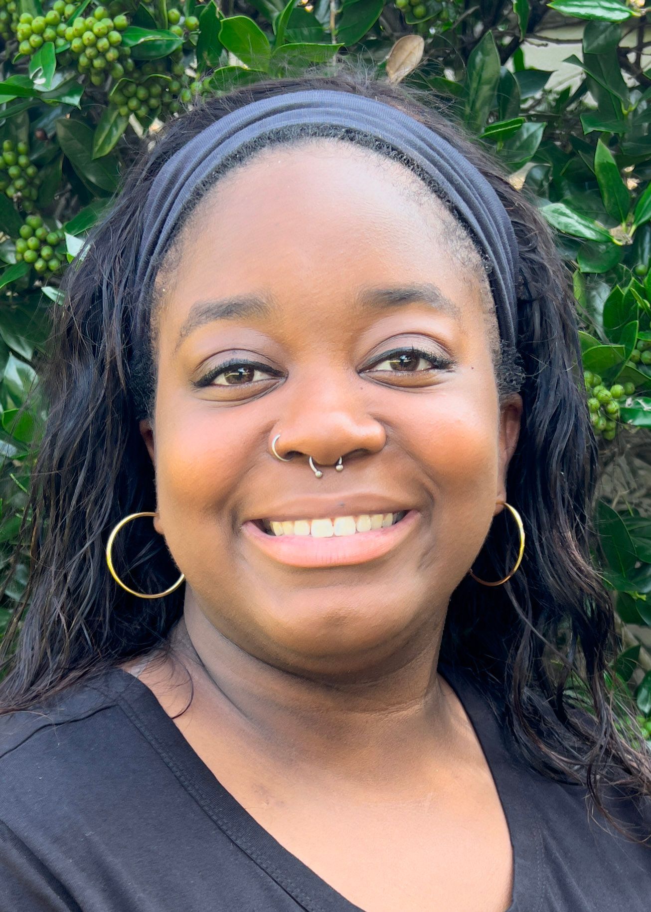 Black woman with long braided hair, nose piercings, and a blue lanyard smiles at the camera in front of leafy green foliage.