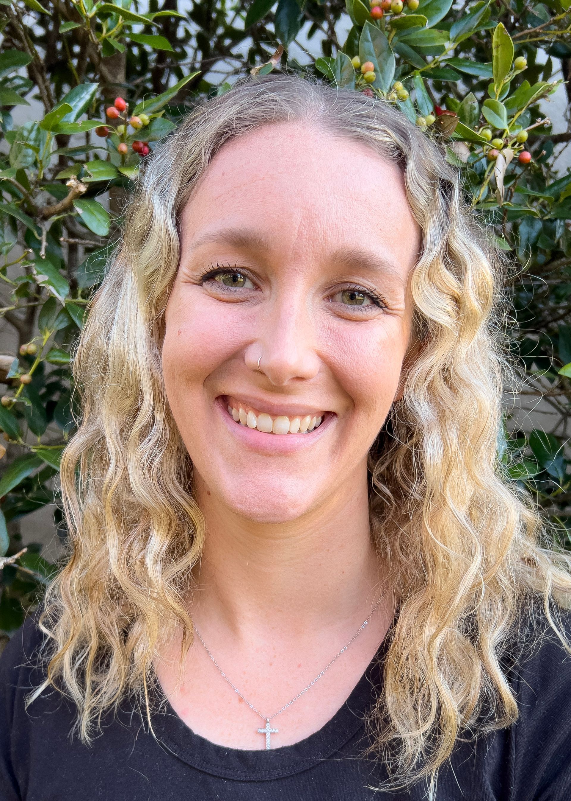 Woman with blonde hair smiling, wearing a black top and gold jewelry, set against a backdrop of green foliage.