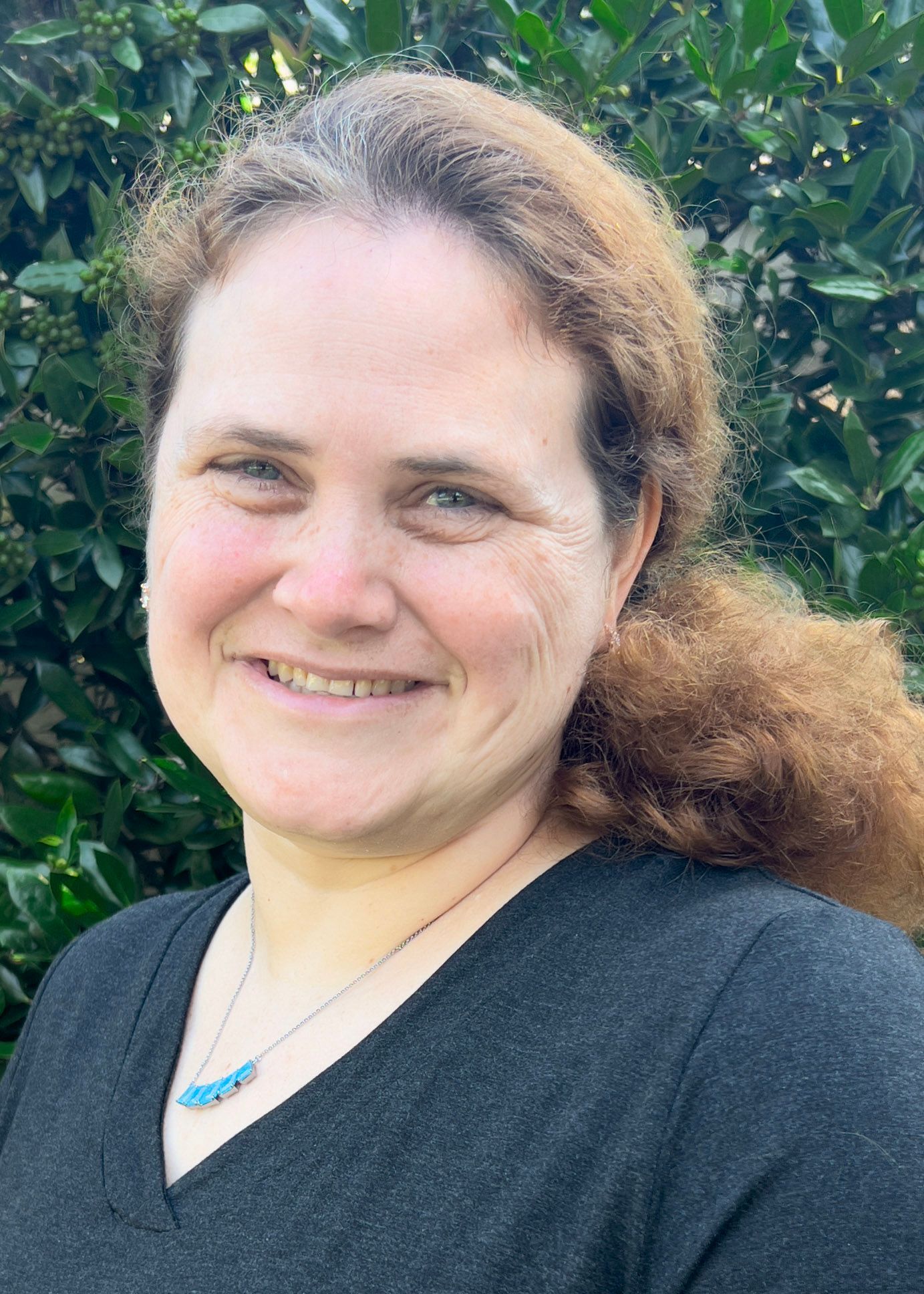 Woman with curly brown hair smiles at the camera, wearing a black sweater in front of a dark blue background.