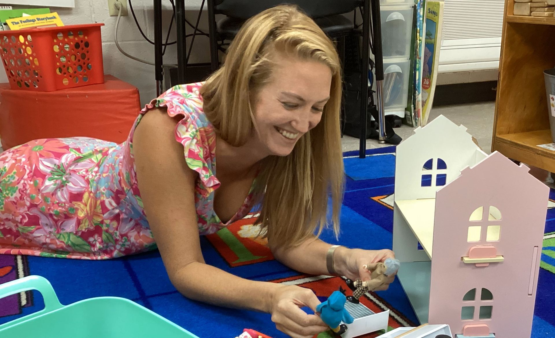 Woman smiles while playing with a dollhouse on a colorful rug in a classroom setting. She wears a floral dress.