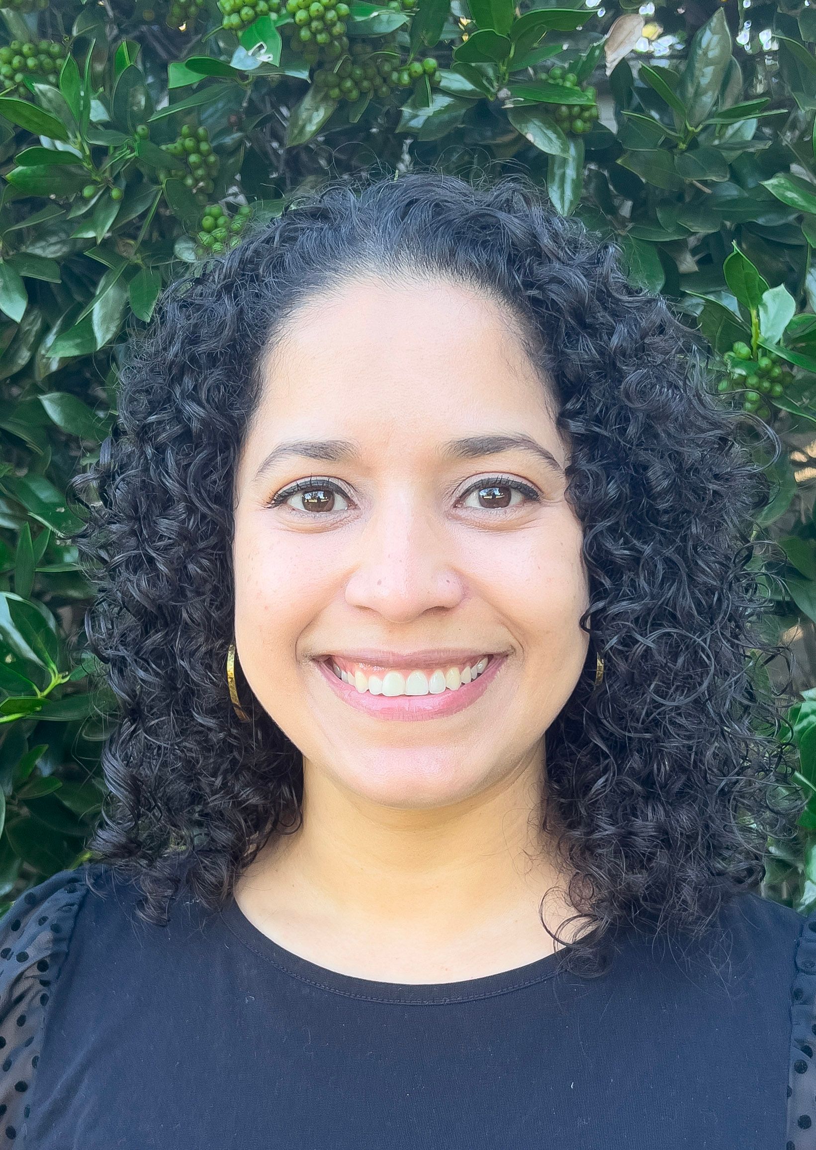 Woman with curly dark hair and a smile wearing a light gray blazer and white top against a blue background.