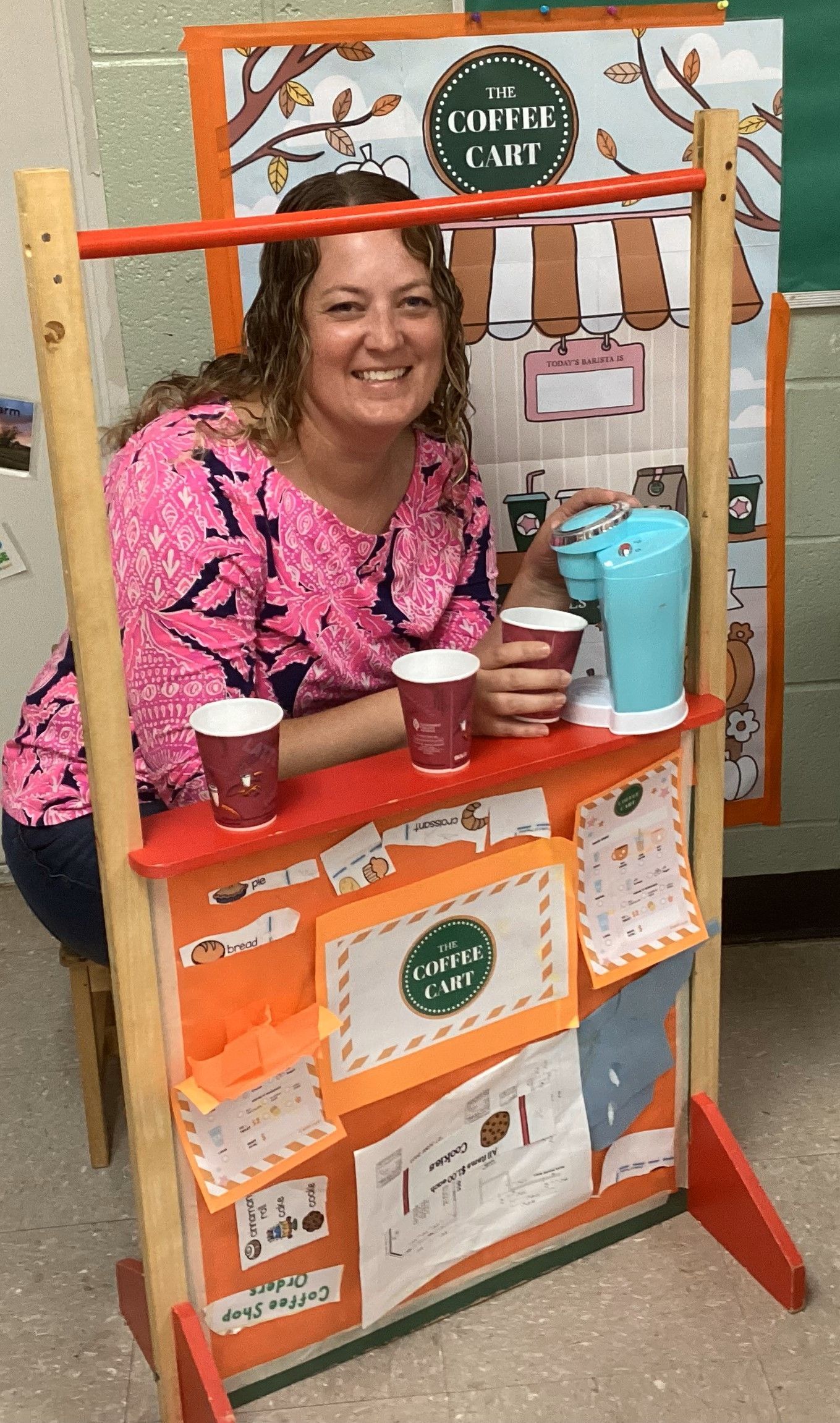 Woman smiling behind a play food cart with red cups and a blue toy drink maker. The cart has artwork and orange accents.