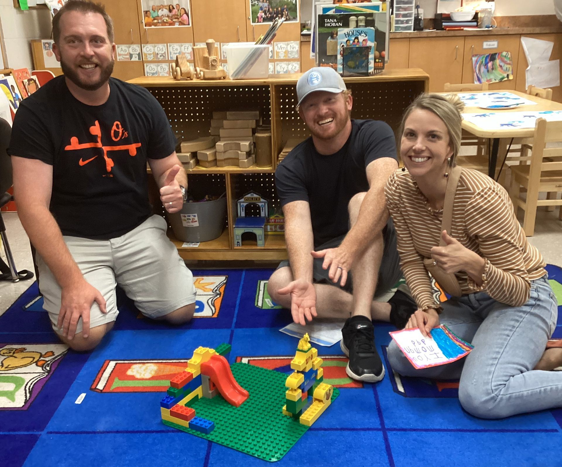 Three people kneel on a blue rug with a Lego structure. They smile and give thumbs-up in a colorful classroom setting.