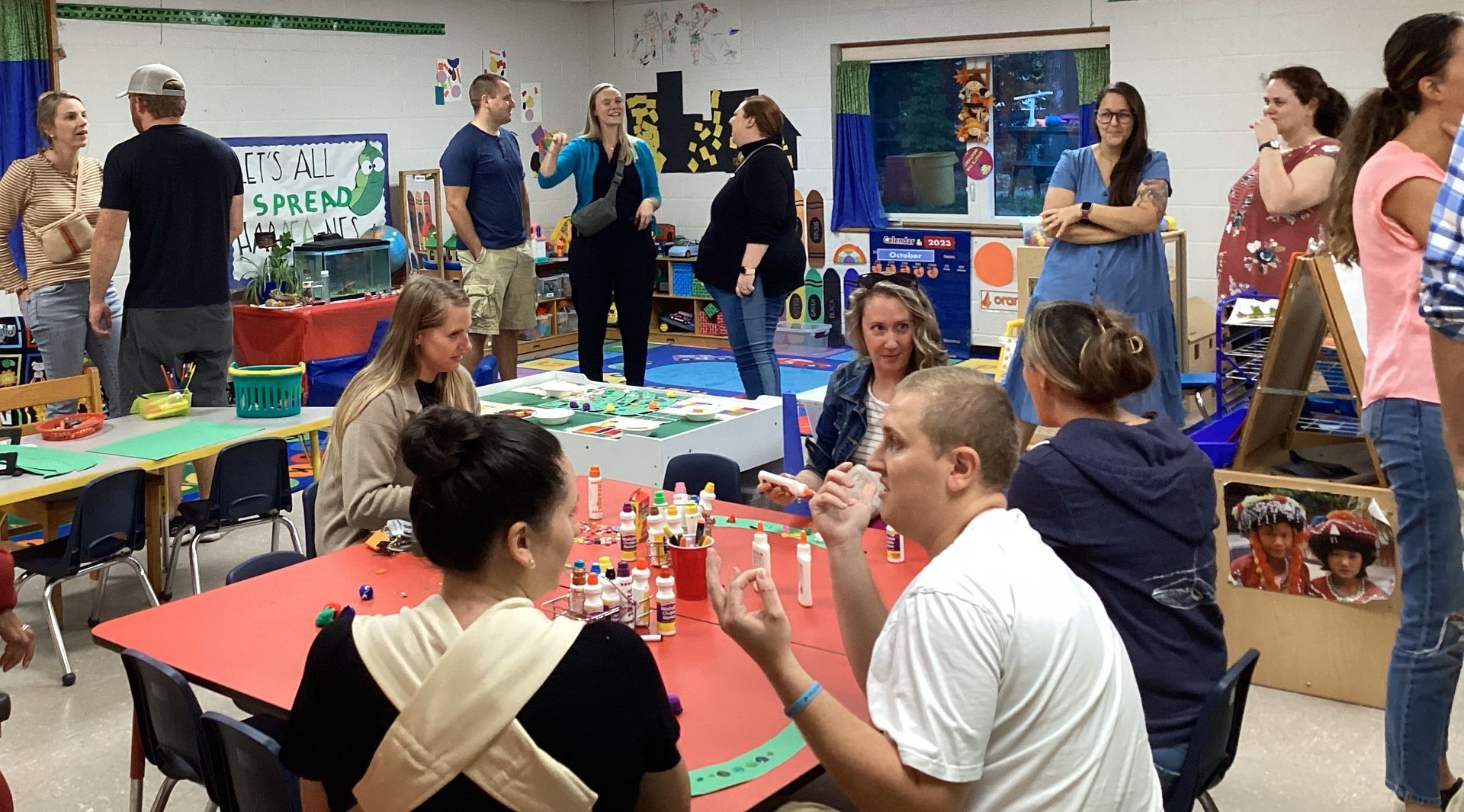 People gathered in a classroom, some seated at red tables playing with Lego. Others are standing and chatting near toys and colorful decorations.