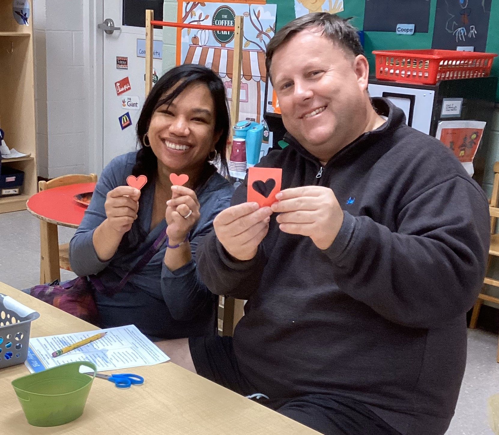 Two smiling people in a classroom hold up paper hearts. One is red, and the other is black on a red card.