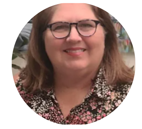 Woman with glasses smiling, wearing a floral top, in front of a painting and a plant.