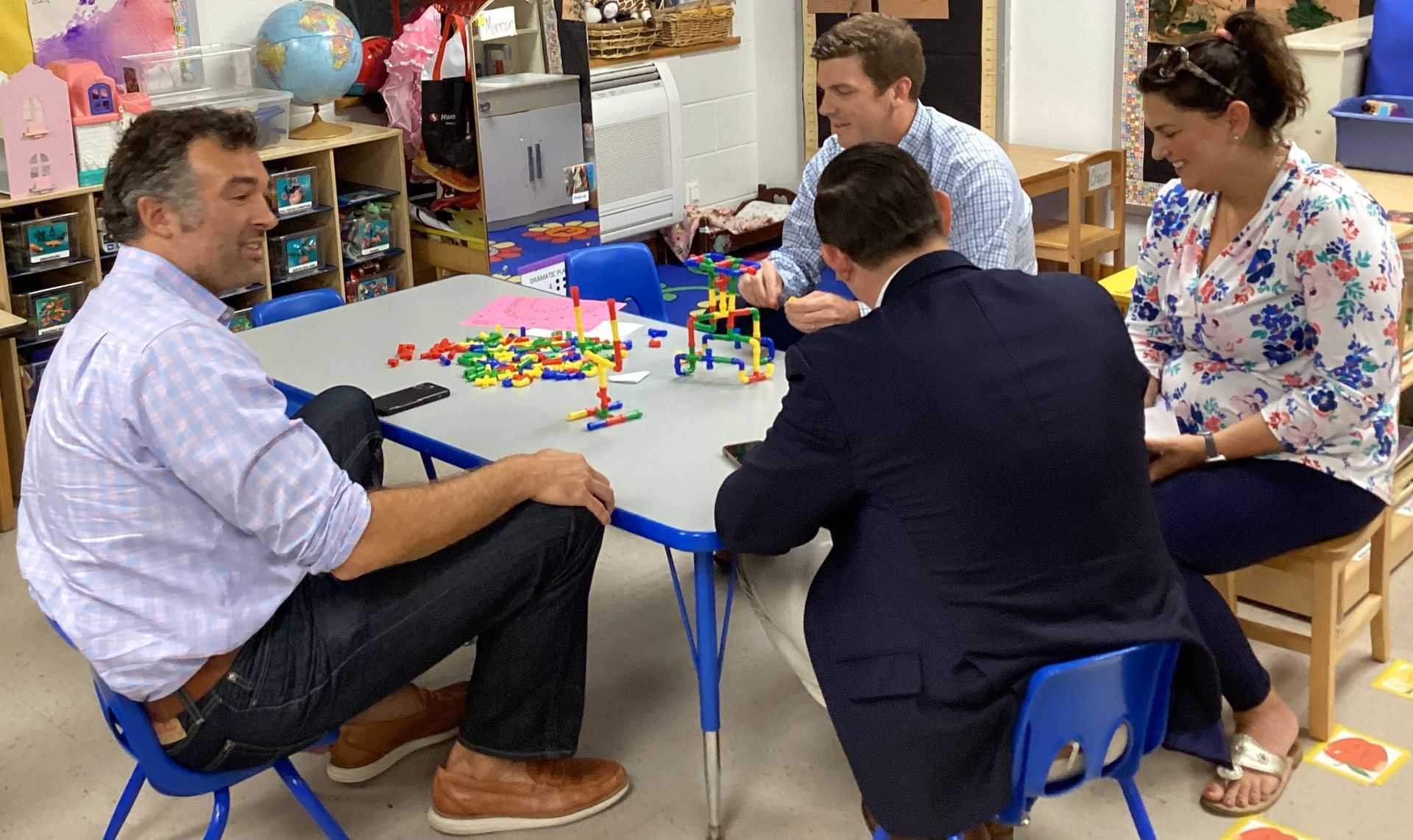 Four adults sit around a small table in a brightly-lit room, playing with colorful building blocks. One man smiles, and another looks intently at the blocks.