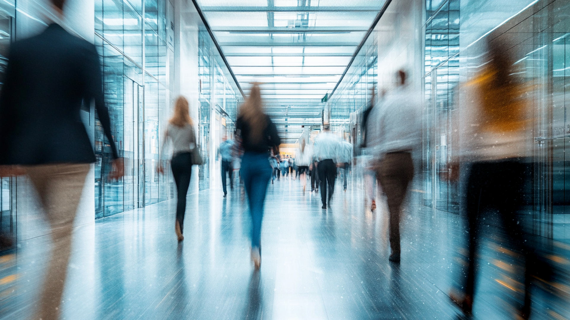Blurred figures walking down a modern hallway with glass walls and ceiling.