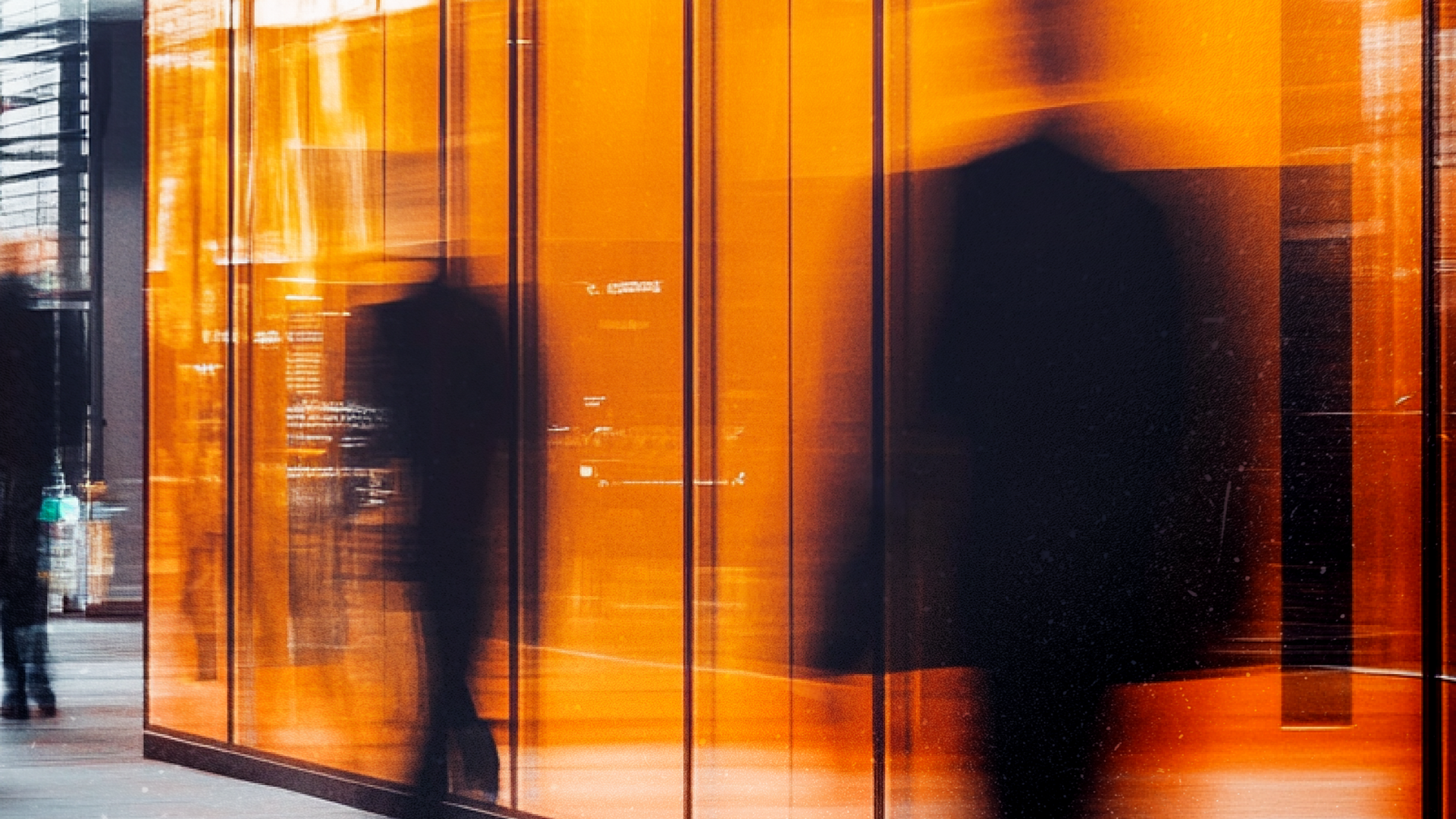 Blurred figures walk by orange glass panels, reflecting interior lights.