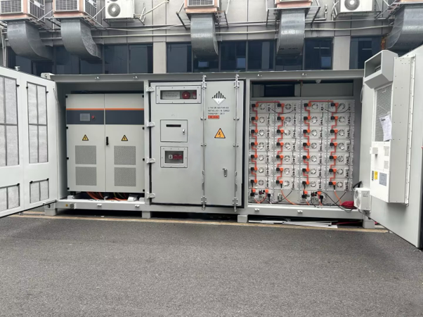 A container with open doors revealing electrical equipment, likely a battery storage system, outdoors near a building.
