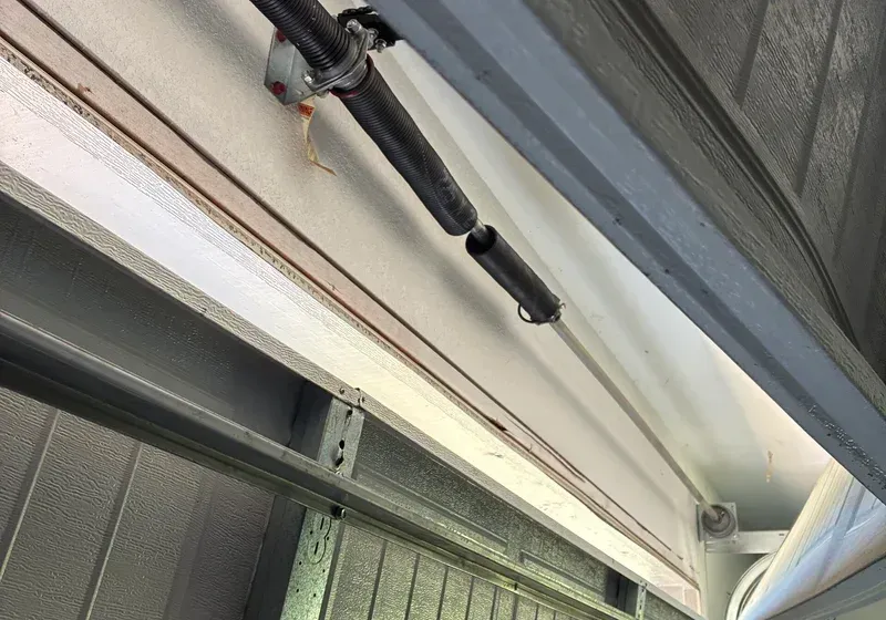 Garage door spring broken, hanging above door frame. White ceiling, gray frame, rusty parts.