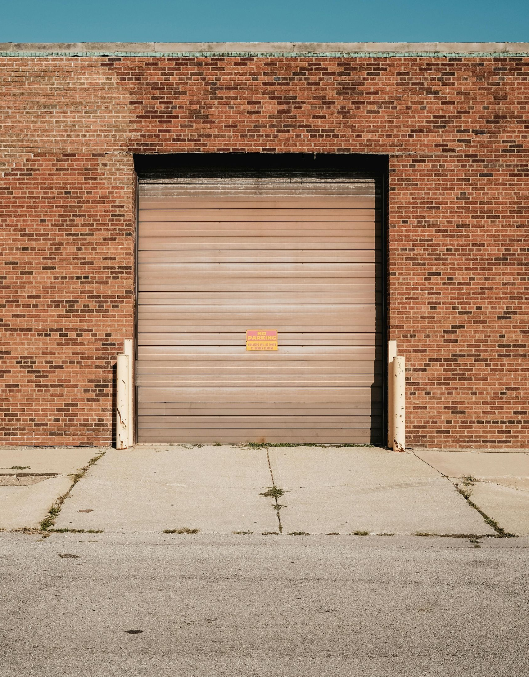 Brown brick building with closed tan loading door; gray concrete pavement below, blue sky above.