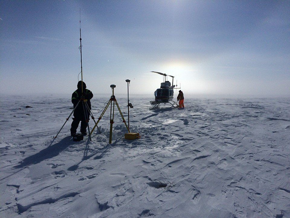 Personne utilisant un équipement d'arpentage près d'un hélicoptère sur un terrain enneigé, soleil éclatant en arrière-plan.