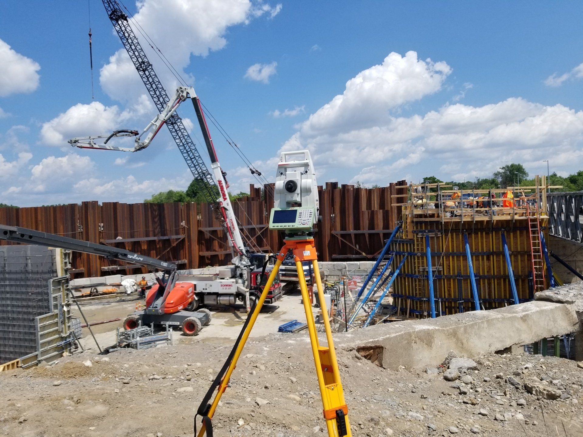 Équipement d'arpentage sur un chantier de construction ; grue, coffrages en béton et mur de soutènement en arrière-plan.