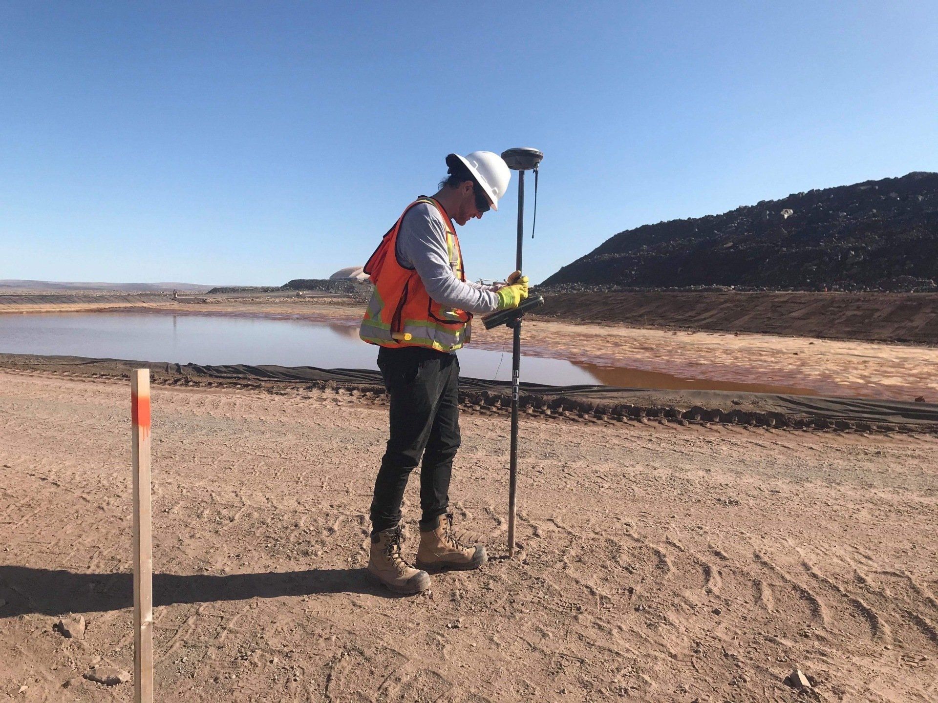 Une personne portant un gilet de sécurité utilise un appareil GPS sur un chemin de terre près de l'eau et des collines.