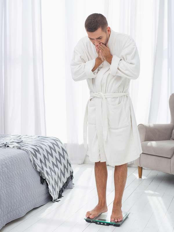 Man in white robe stands on a scale, covering his face in apparent shock of his weight gain due to thyroid dysfunction.