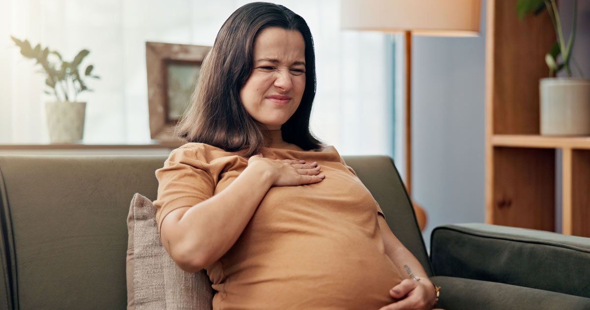 A pregnant woman is sitting on a couch with her hand on her chest needing Acid Reflux Treatments NYC - Dr. John Salerno