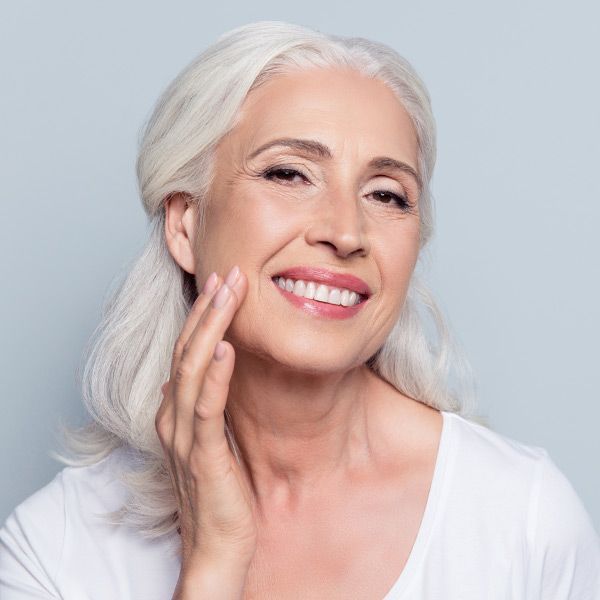 A woman with white hair is smiling and touching her face, happy with her Anti-Aging and Longevity Solutions