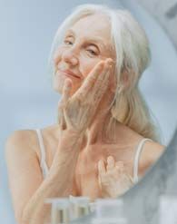Senior woman with white hair applying skincare, looking in the mirror, smiling.
