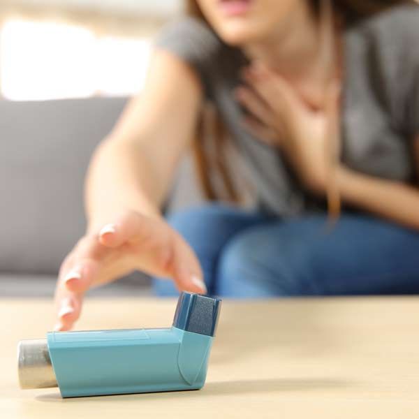 A woman is sitting on a couch using an inhaler, needing Asthma Treatments