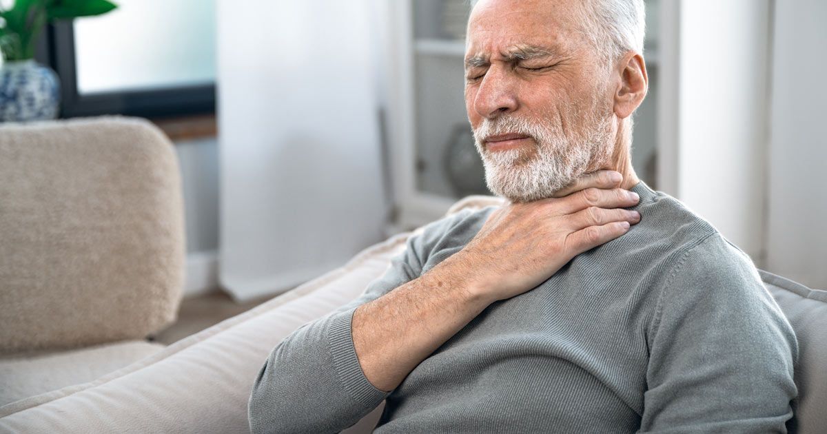 An elderly man is sitting on a couch holding his neck in pain representing Barretts Esophagus Treatments NYC - Dr. John Salerno