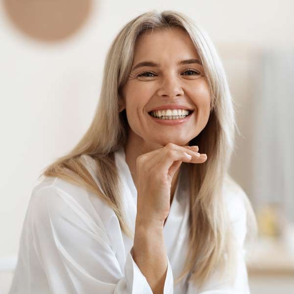A woman in a white shirt is smiling with her hand on her chin after her Beauty Skin IV Treatments