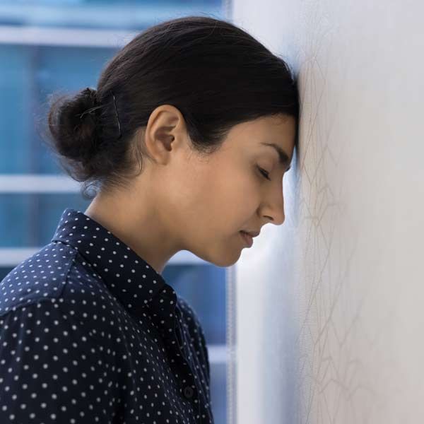 Woman with dark hair in bun presses forehead against a white wall, appearing tired and fatigued. A candidate for biohacking treatments.