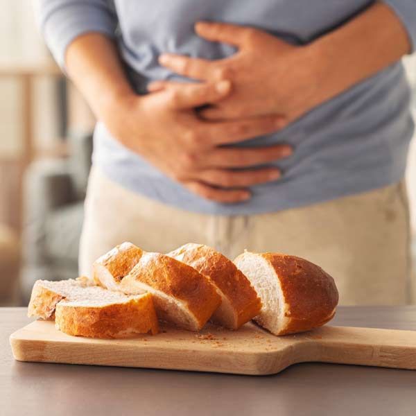 A person is holding their stomach while cutting bread on a cutting board, representing Celiac Disease Treatments