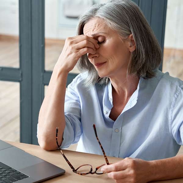 An older woman sits at a desk with a laptop, rubbing her nose after missing her Chronic Fatigue and Viral IV Treatments