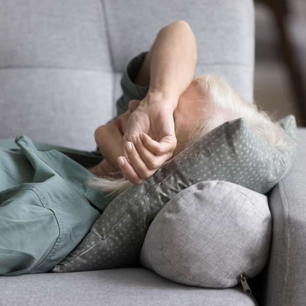 Woman on a gray sofa, face covered with hand, appears to be resting or distressed. Complementary Medical Practitioner in Manhattan, NY, and Connecticut