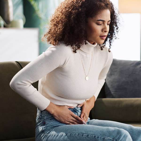 A woman sitting on a couch, clutching her stomach, eyes closed, appearing to be in pain. Indoors, daytime, a prime candidate for Endometriosis Therapy by Salerno Wellness in Manhattan, NY and Connecticut